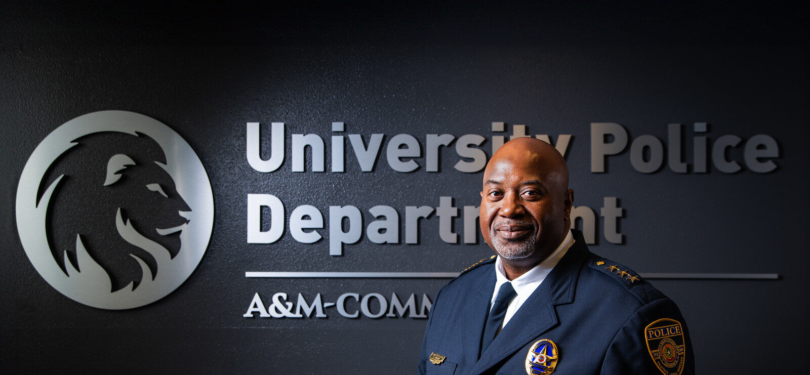 Chief Bryan Vaughn dressed in his dark blue, formal police uniform. He is standing in front of a dark wall with a silver embossed TAMUC lion head logo and the words University Police Department A&M-Commerce.