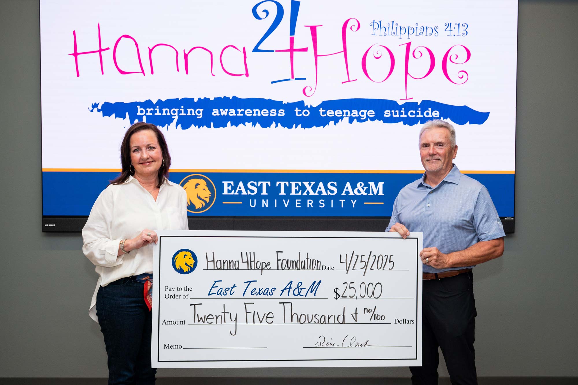 Two people hold a poster-size check, one on each side. Behind them, a screen reads "Hanna4Hope."
