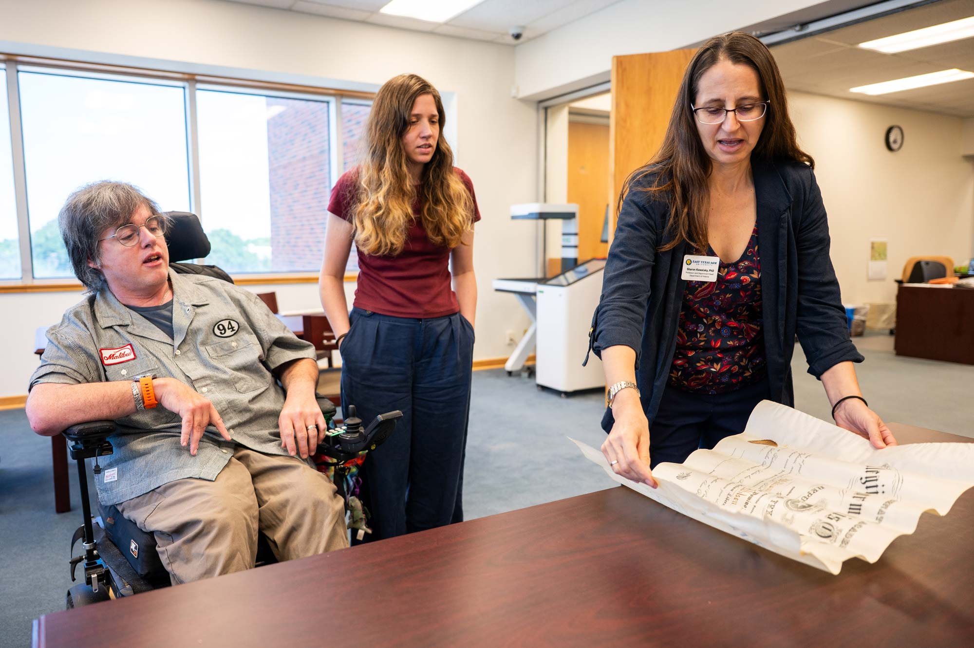 Three people look at an off-white diploma lying on a brown tabletop. The person on the right holds the diploma open.