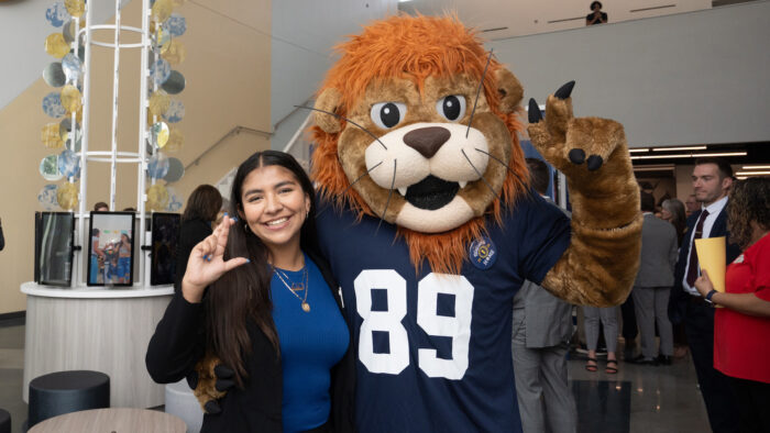 A person in business attire posing for a photo with someone in a lion mascot costume indoors.