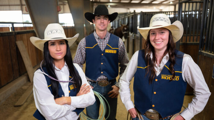 Three people in Western attire pose for a photo inside of a covered dirt arena.