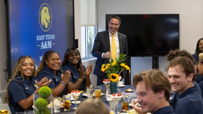 Man stands behind a table and smiles at camera. Three people are seated at the table to his right and around five people are seated at the table to his left.