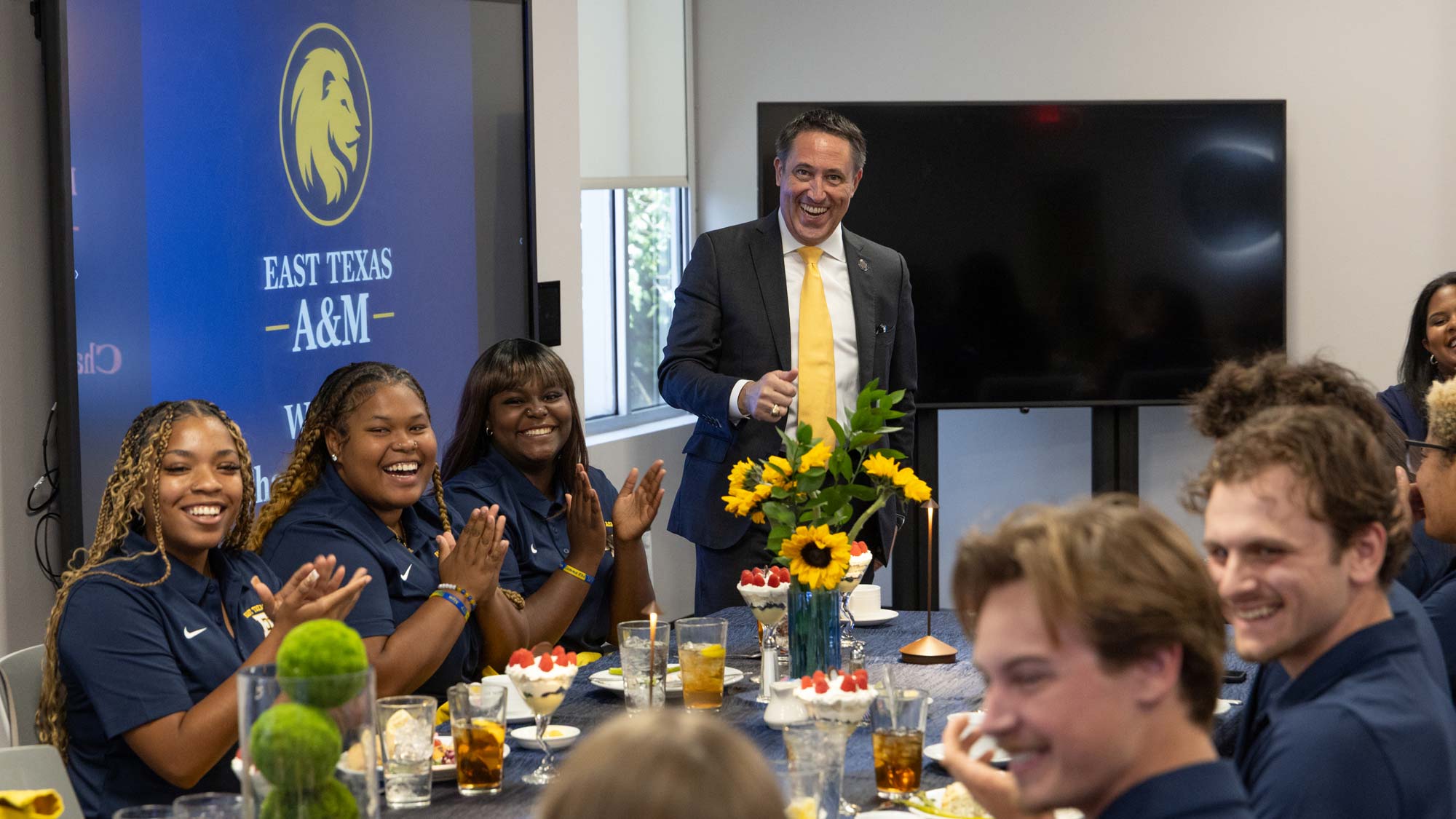 Man stands behind a table and smiles at camera. Three people are seated at the table to his right and around five people are seated at the table to his left.