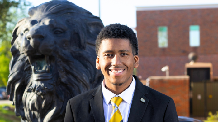 A person in a suit posing for a photo outdoors in front of a Lion statue.