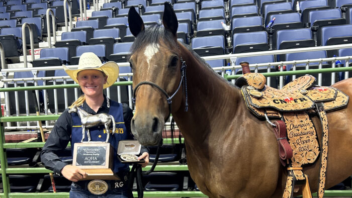 A person in Western attire posing with a trophy next to a horse inside of a rodeo arena.