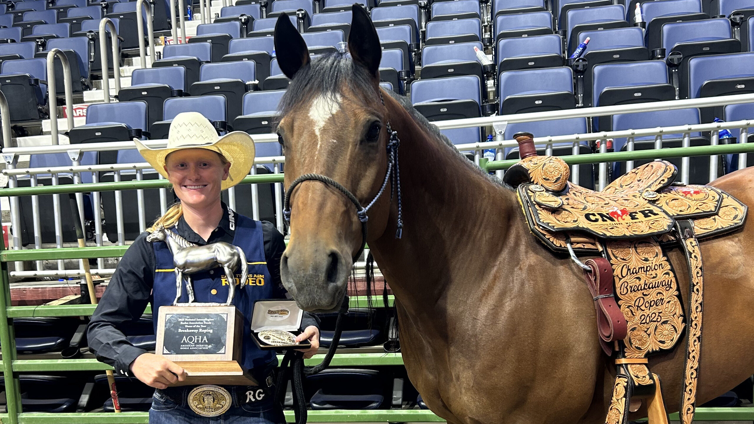 A person in Western attire posing with a trophy next to a horse inside of a rodeo arena.