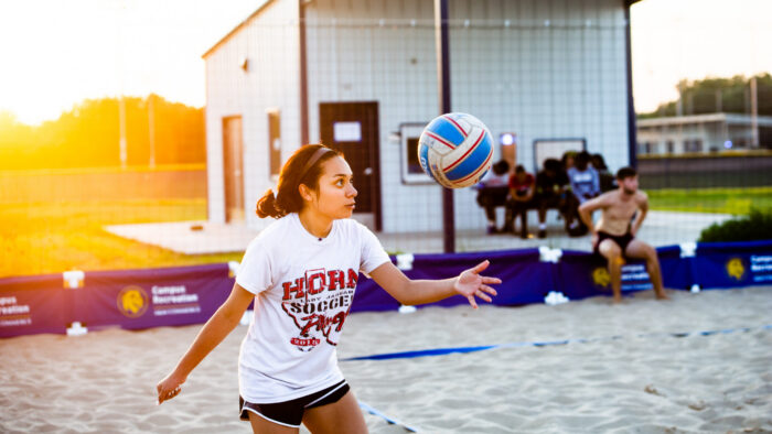 Girl standing in the sand, throwing a volleyball in the air.
