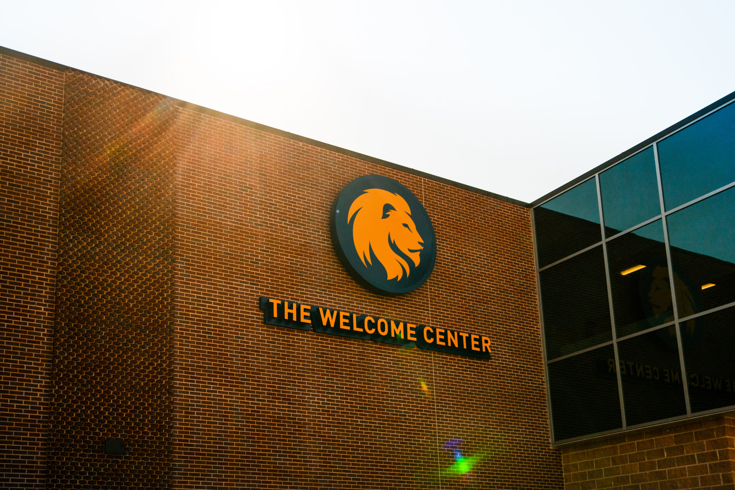 An outdoor brick wall displaying a blue and gold lion logo and a sign that reads "The Welcome Center."