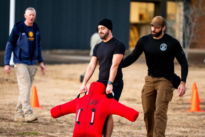 Person pulls red weighted dummy patient while coach stands behind him to support him if needed.