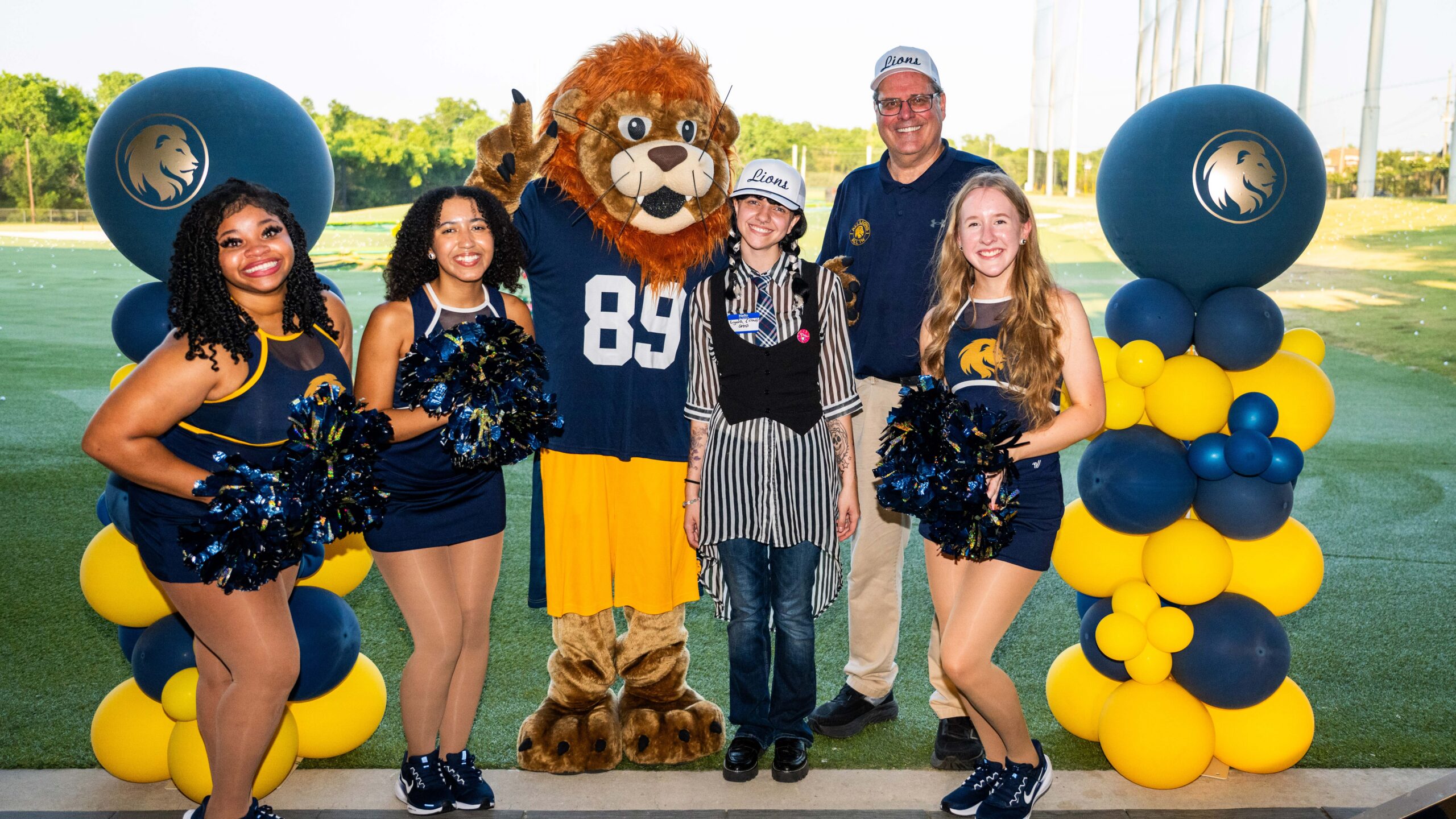 Three cheerleaders standing with a student, a person in a lion costume, and a man with a white hat.