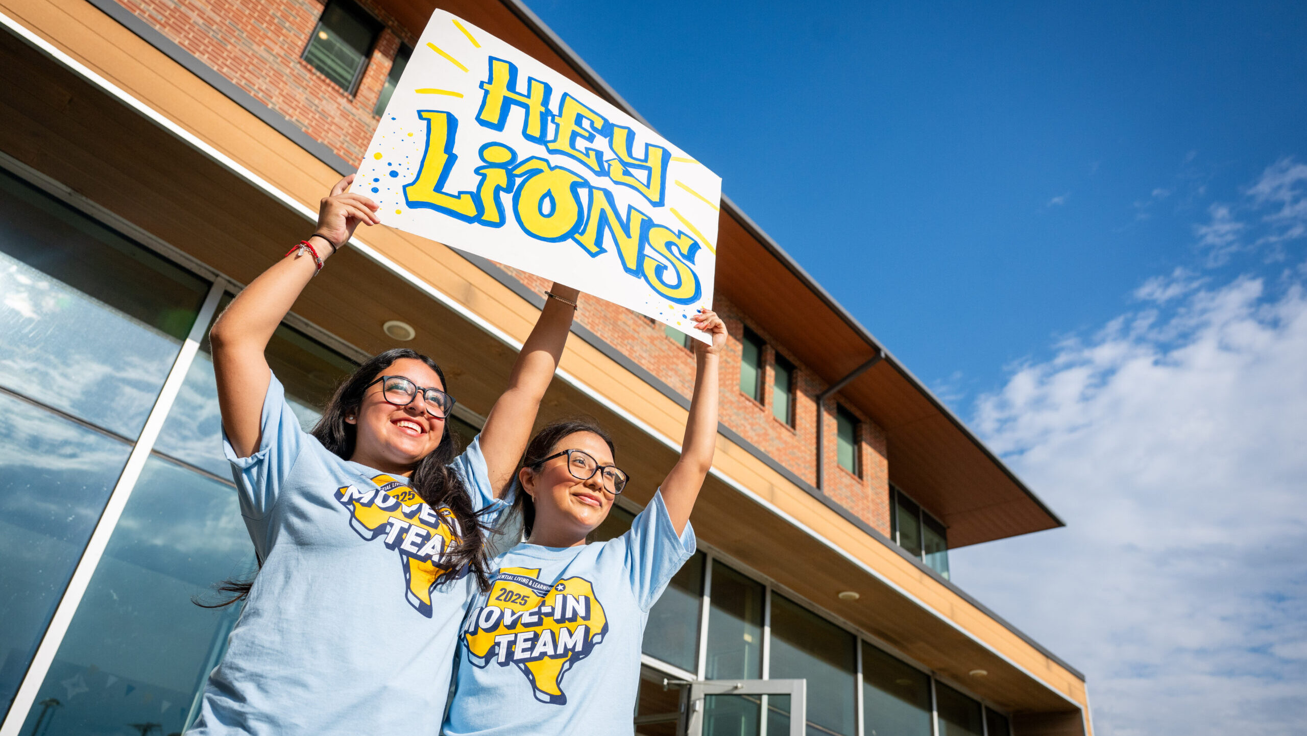 Two people standing outdoors holding a posterboard sign that reads "Hey Lions"