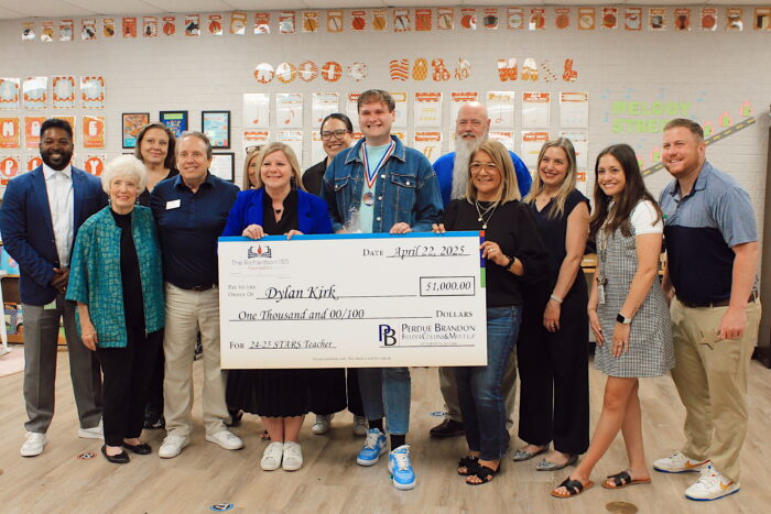Thirteen people standing together and smiling while holding a giant check.