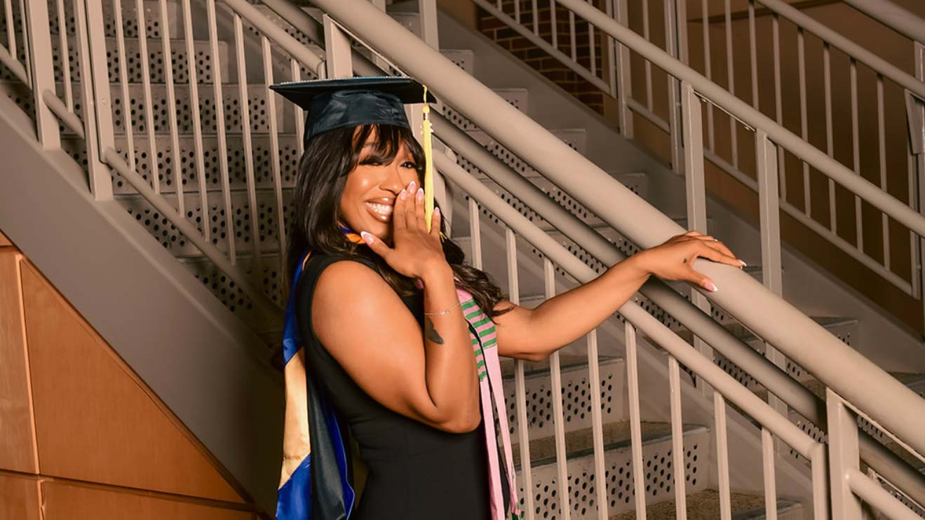 Woman in graduation attire smiling and standing by a set of stairs.