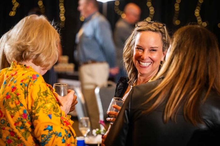 Person at table laughs with two other people whose backs are to the camera. A wine glass is in one of their hands.