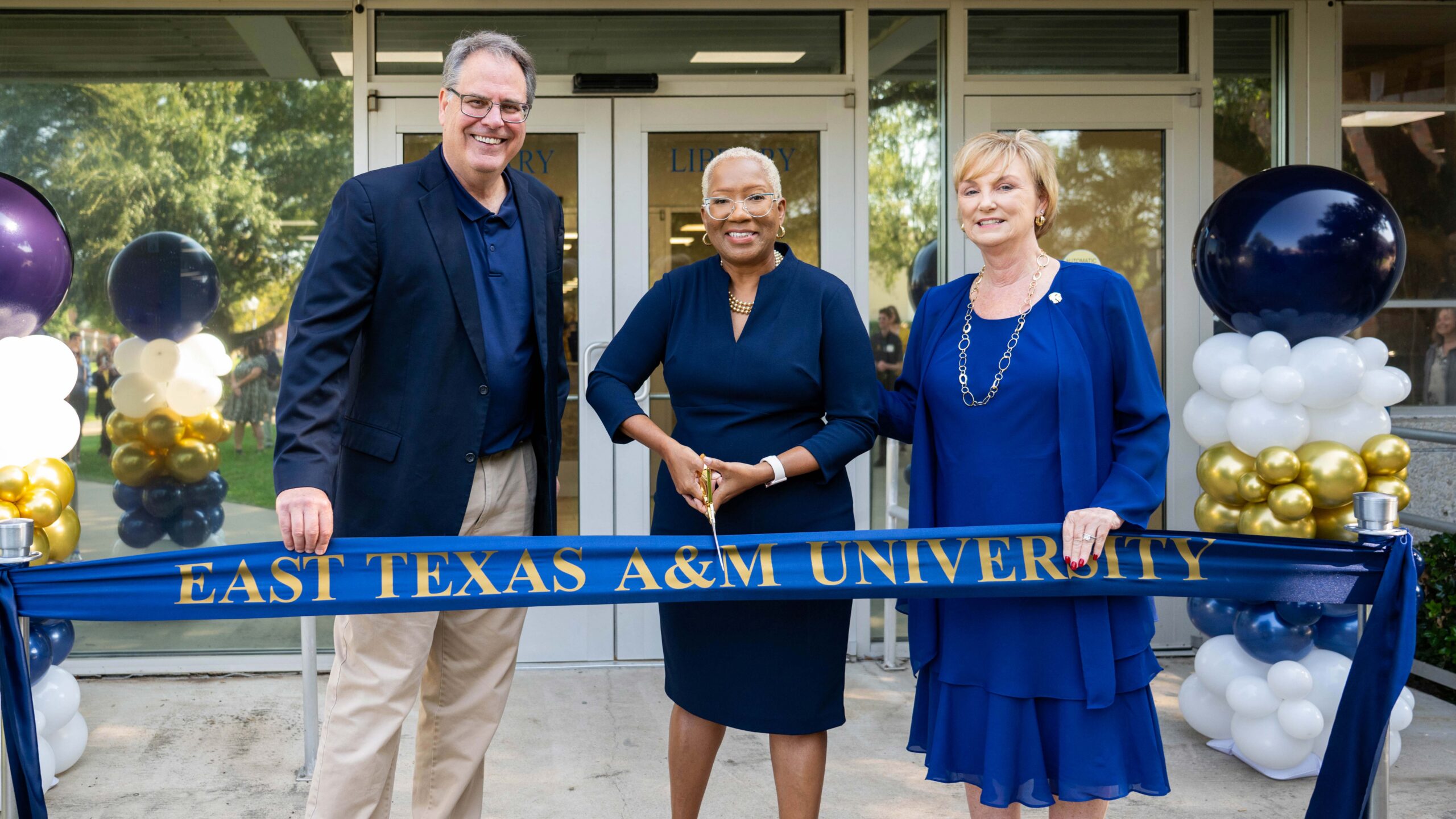 Three people standing outside about to cut a blue ribbon.