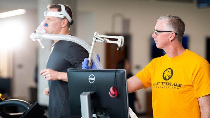 A man watching another man run on a treadmill with a breathing device strapped to his face.