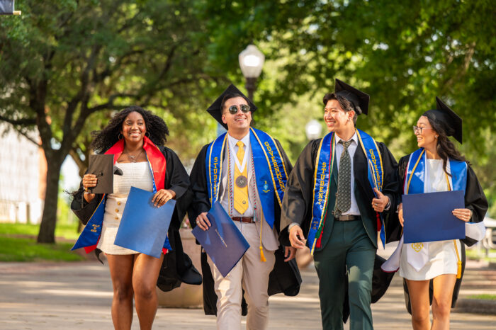 Four students walk down the sidewalk, wearing graduation caps and gowns.