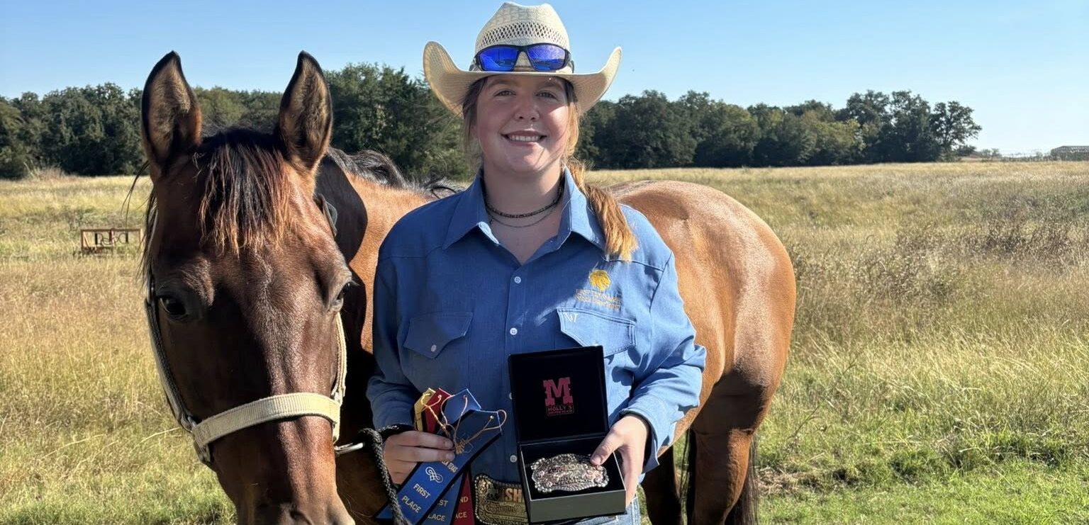 A person in western attire posing with a decorated belt buckle next to a brown horse.