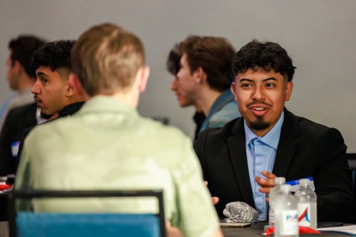 A young man seated at a table talks with another young man.