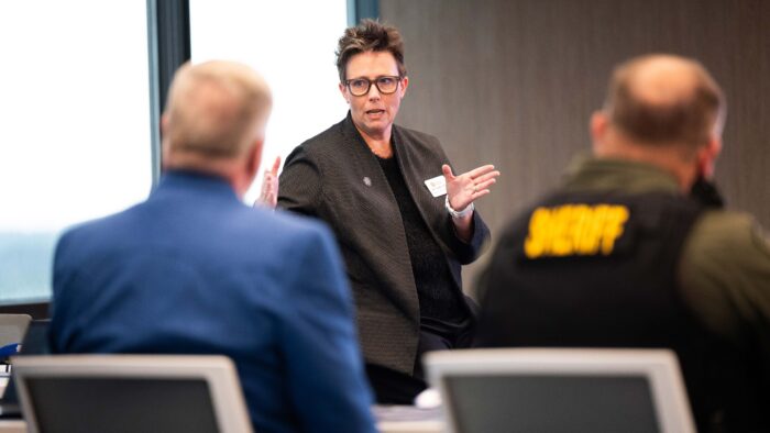 A woman is standing and talking in front of two men seated in chairs.