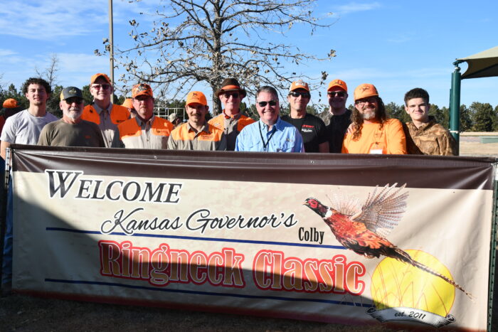 A group of 11 men in various styles of hunting apparel stand behind a large vinyl banner welcoming people to the event.